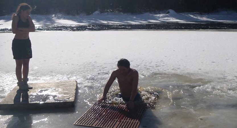a person emerges from a hole cut in a frozen river during a polar plunge. Another stands nearby, watching. 